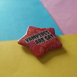 May include: A red star-shaped pin with the text "VAMPIRES ARE GAY" in white. The pin has a pattern of white vampire teeth and other shapes. The background is composed of blue, pink, and yellow.
