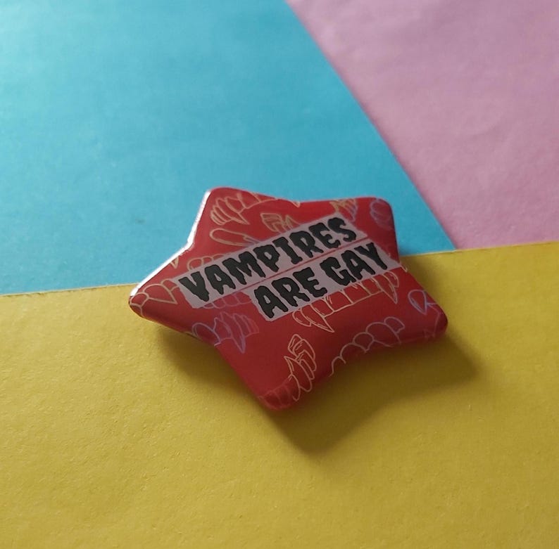 May include: A red star-shaped pin with the text "VAMPIRES ARE GAY" in white. The pin has a pattern of white vampire teeth and other shapes. The background is composed of blue, pink, and yellow.