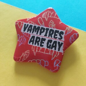 May include: A red star-shaped pin with the text "VAMPIRES ARE GAY" in black and white. The pin features a pattern of vampire teeth in a lighter red color. The pin is on a yellow and blue background.