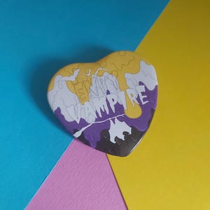 May include: Heart-shaped button with a stylized design. The button features a yellow, white, purple, and black color scheme. The word "VAMPIRE" is printed in white across the center of the button. A bat silhouette is visible.