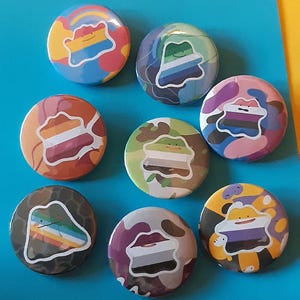 May include: Nine circular pin badges with diverse designs. Each badge displays a stylized shape incorporating pride flag colour schemes, set against backgrounds of camouflage, rainbow, and other patterns. The badges are arranged on a blue surface.