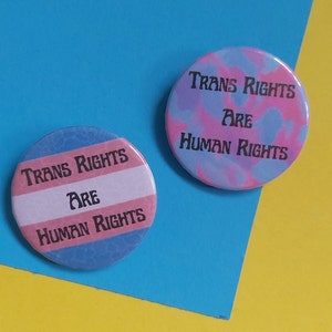 May include: Two round buttons with the text "Trans Rights Are Human Rights" in black lettering. One button has a pink, blue, and white tie-dye pattern. The other button has a blue, pink, and white striped pattern.
