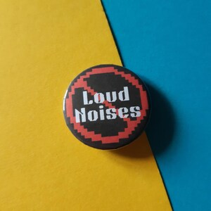 May include: A black and red button with a pixelated design. The button says "Loud Noises" with a red diagonal line through the word "Loud".