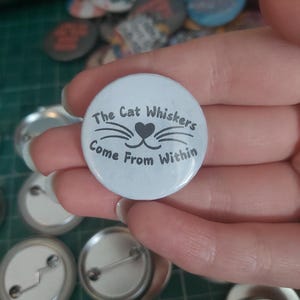 May include: A light blue button with the text "The Cat Whiskers Come From Within" in a black font. The button features a cat face design with whiskers and a heart-shaped nose. Other buttons are visible in the background.