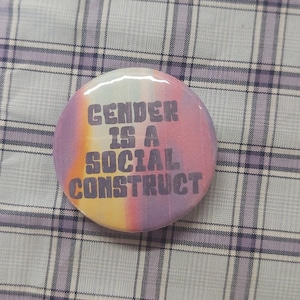 May include: A round button with a colorful tie-dye background in shades of pink, yellow, and blue. The button features the text "GENDER IS A SOCIAL CONSTRUCT" in a dark gray, bold font.