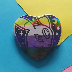 May include: Heart-shaped button with a holographic finish. The design features a white cartoon animal face over a non-binary pride flag background. The button is decorated with stars and a crescent moon.