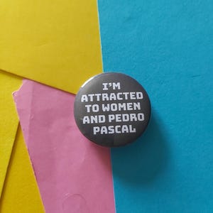May include: A round, dark gray button with white text that reads "I'M ATTRACTED TO WOMEN AND PEDRO PASCAL." The button is centered on a background of yellow, pink, and blue paper.