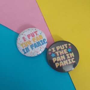 May include: Two circular pin-back buttons. One button is white with a multicolored paint splatter pattern and the text I PUT THE PAN IN PANIC in pink, yellow, and blue. The other button is black with small colorful stars and the same text in yellow.
