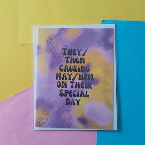 May include: Greeting card with the text "THEY/ THEM CAUSING MAY/HEM ON THEIR SPECIAL DAY" in black font. The background features a purple and yellow watercolor design. The card is in a clear plastic sleeve.