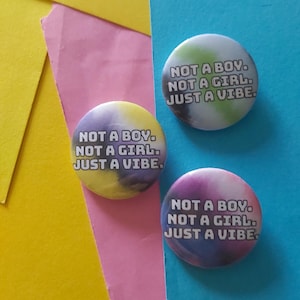Puede incluir: Tres botones redondos con el texto "NOT A BOY. NOT A GIRL. JUST A VIBE." Los botones tienen un diseño colorido y arremolinado en tonos de amarillo, verde y morado. Están colocados sobre un fondo de papel rosa, amarillo y azul.