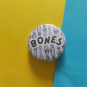 Puede incluir: Un botón redondo blanco con la palabra "BONES" en letras negras en negrita. El fondo del botón presenta un patrón repetido de ilustraciones de huesos en blanco y negro. El botón está sobre un fondo amarillo y azul.