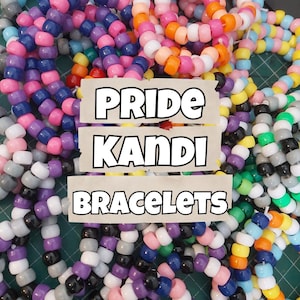 以下が含まれることがあります： カラフルなビーズブレスレットの山をクローズアップした写真で、画像の上に「PRIDE KANDI BRACELETS」というテキストが重ねられています。