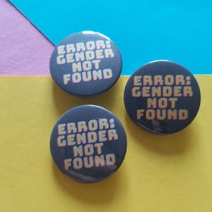 May include: Three black button pins with the text "ERROR: GENDER NOT FOUND" in yellow and pink letters.