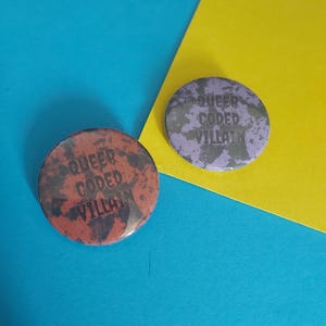 May include: Two round buttons with a black and white splatter paint design. The text "Queer Coded Villain" is printed in black on both buttons. One button is purple and the other is brown.
