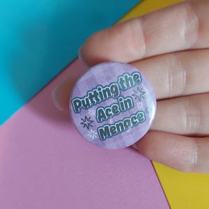 May include: A circular badge in lavender, featuring the text "Putting the Ace in Menace" in white and grey, with star motifs. The badge is held in a hand, set against a colourful backdrop.