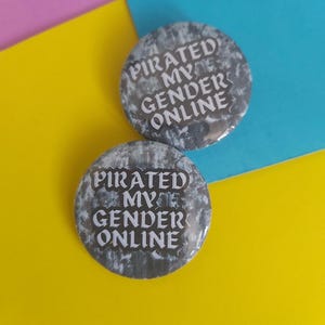 May include: Two round buttons with the text "PIRATED MY GENDER ONLINE" in white lettering. The buttons have a textured, gray and white background. The background is a mix of yellow, blue, and pink.