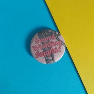 May include: A circular badge featuring the text "Sword Wielding Sapphic" in pink. The badge has a light pink background with a black and grey illustration. The background is blue and yellow.