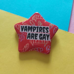 Puede incluir: Un botón rojo en forma de estrella con el texto "VAMPIRES ARE GAY" en letras negras. El botón tiene un patrón de colmillos de vampiro en un color rojo más claro. El fondo es una mezcla de amarillo y azul.