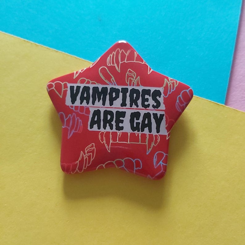 May include: A red star-shaped button with the text "VAMPIRES ARE GAY" in black letters. The button has a pattern of vampire fangs in a lighter red color. The background is a mix of yellow and blue.