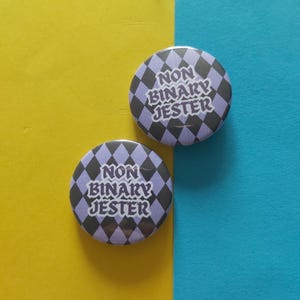 Könnte beinhalten: Zwei runde Buttons mit dem Text "NON BINARY JESTER" in Weiß, vor einem lila-schwarzen Rautenmuster. Die Buttons befinden sich auf einem gelb-blauen Hintergrund.