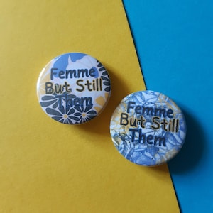 May include: Two round buttons with a blue and yellow floral pattern. The text on the buttons reads "Femme But Still Them".