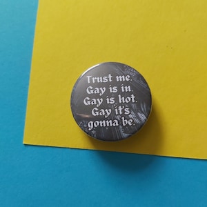 Peut inclure: Un bouton argenté avec une inscription blanche qui dit "Trust me. Gay is in. Gay is hot. Gay it's gonna be."