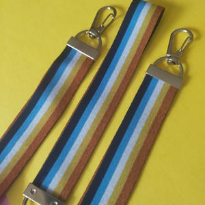 Könnte beinhalten: Drei gestreifte Schlüsselbänder mit silberfarbenen Metallverschlüssen. Jedes Schlüsselband hat ein sich wiederholendes Muster aus schwarzen, blauen, weißen, gelben und braunen Streifen. Die Schlüsselbänder werden vor einem gelben Hintergrund gezeigt.