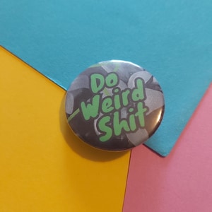 Puede incluir: Un botón redondo con el texto "Do Weird Shit" en letras verdes. El fondo del botón es una mezcla de tonos grises y plateados. El botón está sobre un fondo de papel amarillo, azul y rosa.