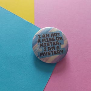 May include: A round button with the text "I AM NOT A MISS OR MISTER. I AM A MYSTERY" in white letters. The button has a blue and purple watercolor design. The background is a mix of blue, yellow, and pink.