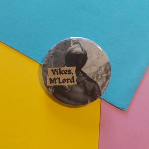 May include: A circular badge featuring a monochrome image of a knight in armour. The text "Vikes, M'Lord" is displayed on a tan label. The badge is set against a backdrop of blue, yellow, and pink paper.