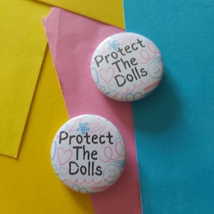 Peut inclure: Deux badges blancs avec le texte "Protect The Dolls" en noir. Les badges présentent un motif floral, des cœurs et des visages souriants en rose et bleu. Le fond est un mélange de jaune, rose et bleu.