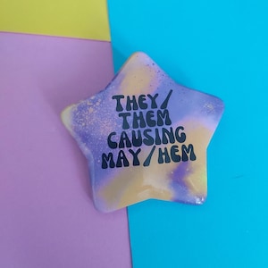 May include: A star-shaped badge with a yellow, purple, and blue colour gradient. The badge has the text "THEY/ THEM CAUSING MAY/HEM" in black, bold letters. The badge is set against a backdrop of yellow, purple, and blue.