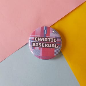 May include: A circular badge featuring the words "CHAOTIC BISEXUAL" in white text. The badge has a pink, red, and blue plaid design. The background is a combination of pink, blue, and yellow.