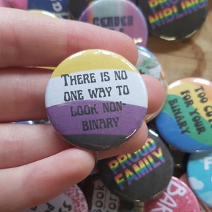 May include: A yellow, white, purple, and black button with the text "There is no one way to look non-binary".