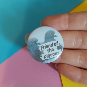 May include: A circular badge with a light blue background. It features two grey pigeons and the text "Friend of the pigeons." The badge is held by a hand against a backdrop of pink, blue, and yellow.