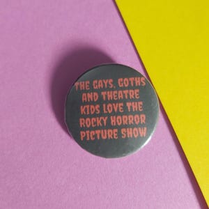 以下が含まれることがあります： 「THE GAYS, GOTHS AND THEATRE KIDS LOVE THE ROCKY HORROR PICTURE SHOW」と赤い文字で書かれた丸い黒いボタン。ピンクと黄色の背景に配置されています。
