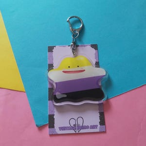 May include: A clear acrylic keychain featuring a cartoon character in yellow, white, purple, and black, with a silver keyring. The keychain is attached to a purple and white striped card with the text "VINTAGE MEMO ART".