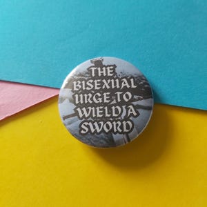 May include: A round button with the text "THE BISEXUAL URGE TO WIELD A SWORD" in white lettering against a blue and gray background. The button is set against a backdrop of blue, pink, and yellow.