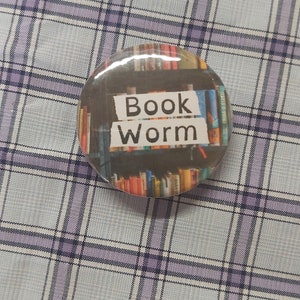 以下が含まれることがあります： 本棚を背景にした、光沢のある丸いボタン。テキスト「Book Worm」は白の太字で表示されています。ボタンは、薄紫と白のチェック柄の背景に配置されています。