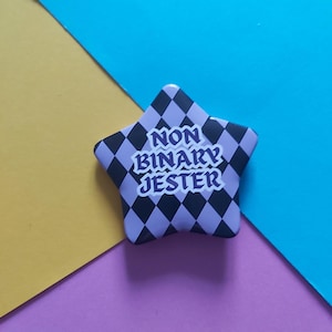 May include: A star-shaped pin with a black and lavender diamond pattern. The pin features the text "NON BINARY JESTER" in white and purple. The background is a mix of yellow, purple, and blue.