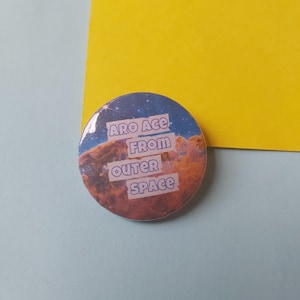 May include: A round button with a space-themed background. The button has text that reads "Aro Ace from outer space" in a purple font.
