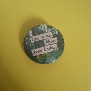 Puede incluir: Un botón redondo con un fondo de escena de bosque. El botón tiene texto que dice "Talk to me About Queer Ecology".