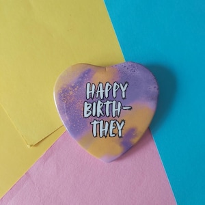 Puede incluir: Botón en forma de corazón con el texto "HAPPY BIRTH-THEY" en blanco, sobre un fondo de acuarela morado y naranja. El botón está sobre un fondo colorido de amarillo, rosa y azul.
