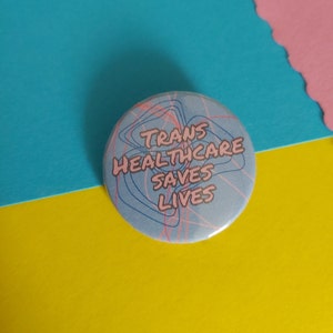 May include: A blue button with pink text that reads "Trans Healthcare Saves Lives" on a blue and pink background.