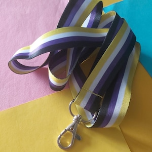 May include: A non-binary pride lanyard with a silver-colored clasp. The lanyard features a repeating pattern of yellow, white, purple, and black stripes. The ribbon is folded and curved, creating a dynamic visual.