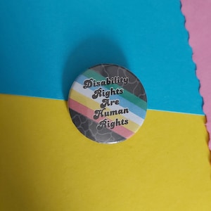 May include: A round button with a black and white background and a rainbow stripe design. The text on the button reads "Disability Rights Are Human Rights".