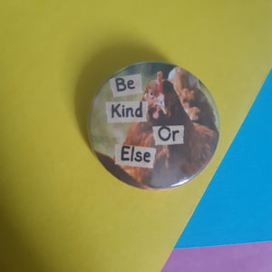 May include: A round button featuring a picture of two chickens. The button displays the text "Be Kind Or Else" in black lettering on white paper.