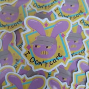 May include: A collection of stickers featuring a purple bunny wearing a gray cap. The bunny has an angry expression and the text "I Don't Care" is written on the sticker. The background is yellow and blue.