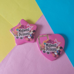 May include: Two pink Midwest Princess themed badges, one star-shaped and one heart-shaped. Both badges feature the text "Midwest Princess" with floral and record designs. The background is a mix of yellow, pink, and blue.