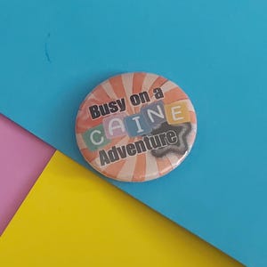 以下が含まれることがあります： 「Busy on a CAINE Adventure」の文字が入った丸いボタン。カラフルなデザインで、レトロなオレンジと白のストライプの背景があります。文字は様々な色とスタイルです。